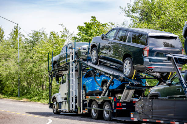 A car carrier truck loaded with multiple vehicles drives along a tree-lined road on a sunny day, providing reliable MI Car Shipping Flint services.