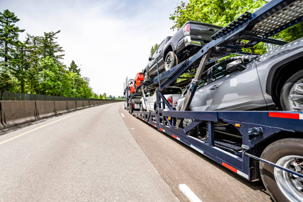 Classic American industrial bonnet car hauler white big rig semi truck tractor transporting cars on the hydraulic two level modular semi trailer driving on the wide turning highway road